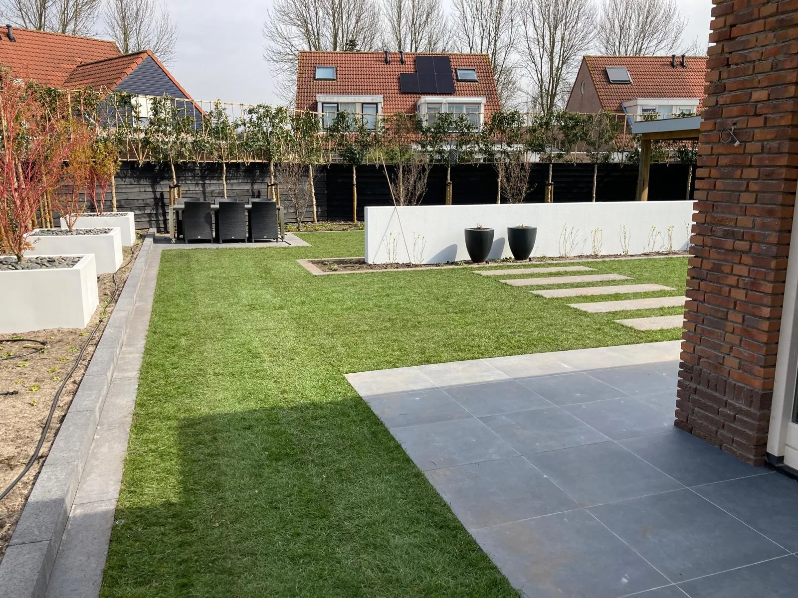 Achtertuin met strak gazon, witte plantenbakken en een keramisch terras